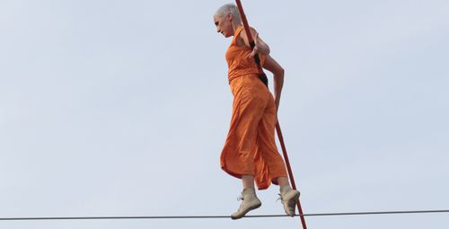 Eine orange gekleidete Person mit rasiertem Kopf balanciert auf einem Seil vor dem Hintergrund des Himmels